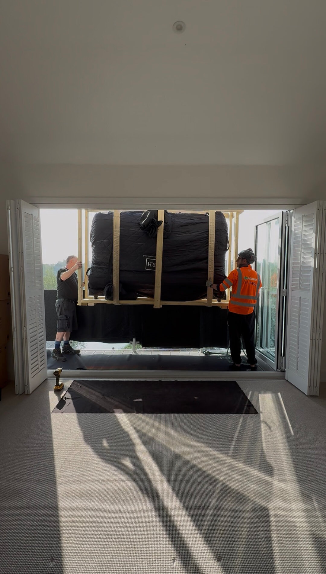RH team guiding a crated, wrapped bed through open balcony doors into a top-floor room